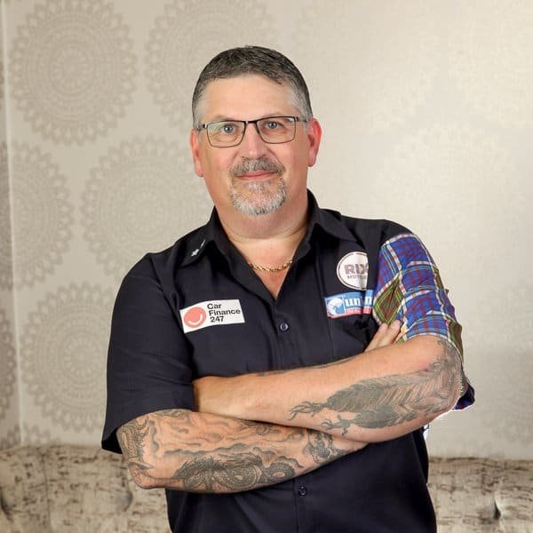 A middle-aged man with glasses, short gray hair, and arm tattoos stands with arms crossed. He wears a black shirt with various patches and a plaid patterned sleeve. The background features a beige wall with circular designs.