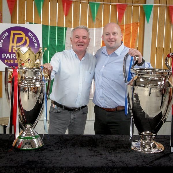 Two men stand side by side, smiling with their arms around each other, behind a table displaying two large sports trophies. A sign for "Parks Pub & Club" and a tricolor flag hang in the background.