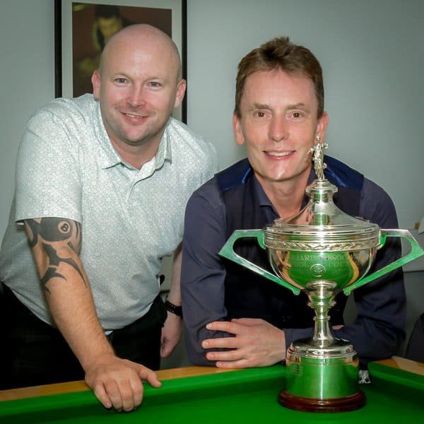 Two men pose indoors behind a large silver trophy on a snooker table. One man is standing with a tattoo on his forearm, while the other leans forward, both smiling at the camera. A framed picture hangs on the wall behind them.