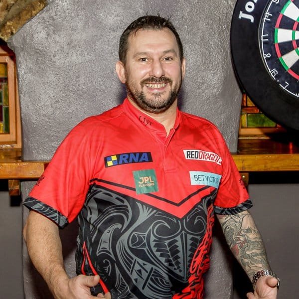 A man wearing a red and black patterned shirt with sponsor logos stands indoors, smiling, with a dartboard visible in the background on the right side.