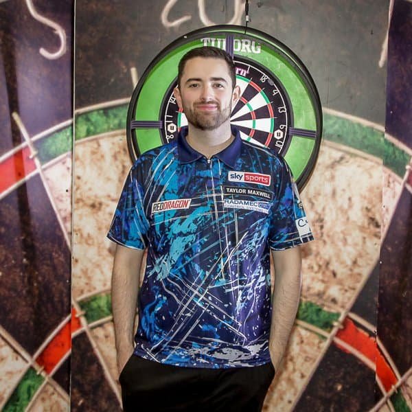 A man in a blue patterned dart shirt stands in front of a dartboard, hands in his pockets, looking at the camera and smiling slightly. The dartboard and background have green, red, and black colors.
