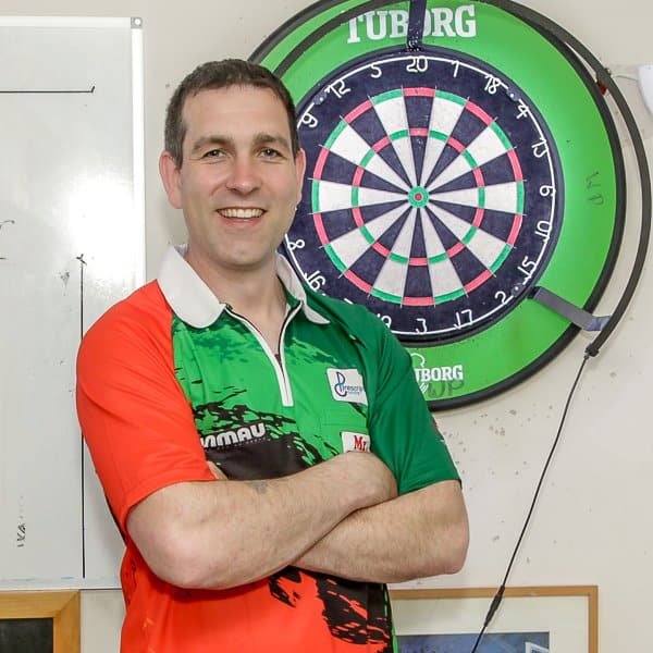 A man wearing a colorful dart shirt stands with arms crossed and smiles in front of a dartboard mounted on a wall. A whiteboard is partially visible on the left side of the image.