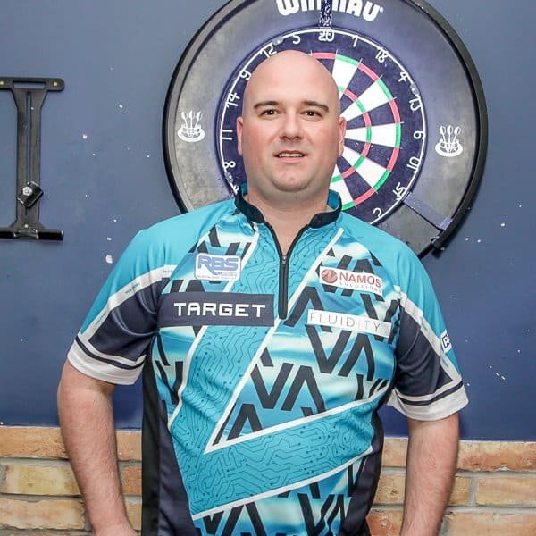 A man in a turquoise and dark blue dart shirt stands in front of a dartboard mounted on a dark blue wall. He is looking at the camera with a neutral expression.
