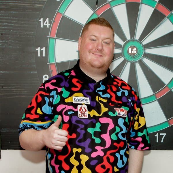 A man stands in front of a dartboard, giving a thumbs up. He is wearing a colorful, abstract-patterned shirt with various sponsor logos. The dartboard is mounted on a dark wall behind him.