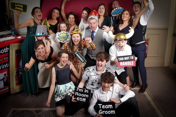 A group of people pose indoors at a photo booth, some wearing hats and holding playful signs with phrases like “Too Hot to Handle,” “I’m in Room 65!!!,” and “Team Groom.” Everyone appears to be celebrating.