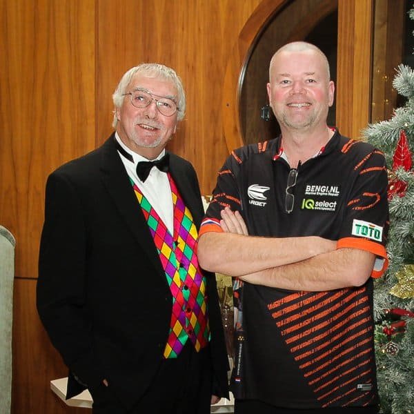 Two men stand indoors beside a decorated Christmas tree. One wears a black tuxedo with a colorful diamond-patterned vest and bow tie; the other wears a black sports shirt with orange accents and logos. Both are smiling.