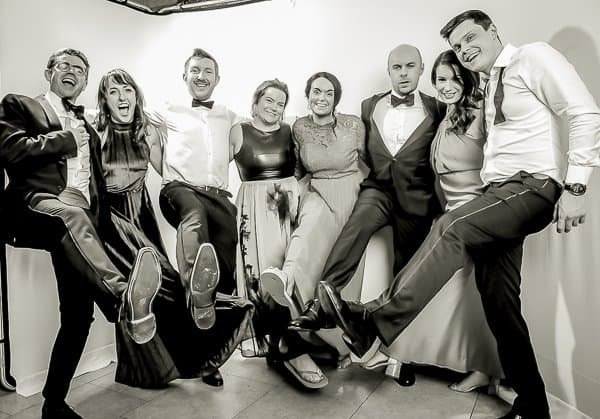A group of four men and four women dressed formally pose indoors, standing side by side with arms linked and one leg raised in front of them, smiling at the camera. The image is in black and white.
