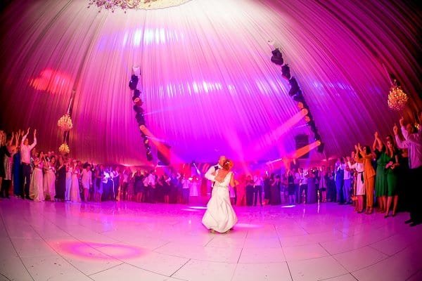A couple dances in the center of a spacious, brightly lit ballroom with a pink and purple glow, while a crowd of guests stands around them, cheering and clapping.